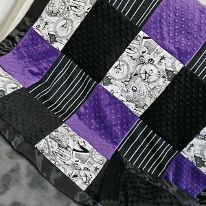 May include: A patchwork blanket featuring squares of black, white, and purple fabrics. The design includes black and white striped fabric, black textured fabric, and purple dimpled fabric. The blanket is trimmed with a black satin border.