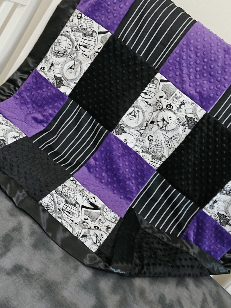 May include: A patchwork blanket featuring squares of black, white, and purple fabrics. The design includes black and white striped fabric, black textured fabric, and purple dimpled fabric. The blanket is trimmed with a black satin border.