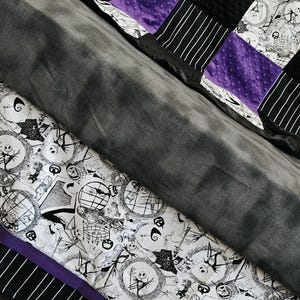 May include: A patchwork quilt featuring a black and white print with Jack Skellington characters, black and white striped panels, and purple accents. The quilt has a soft, gray minky fabric on the back.