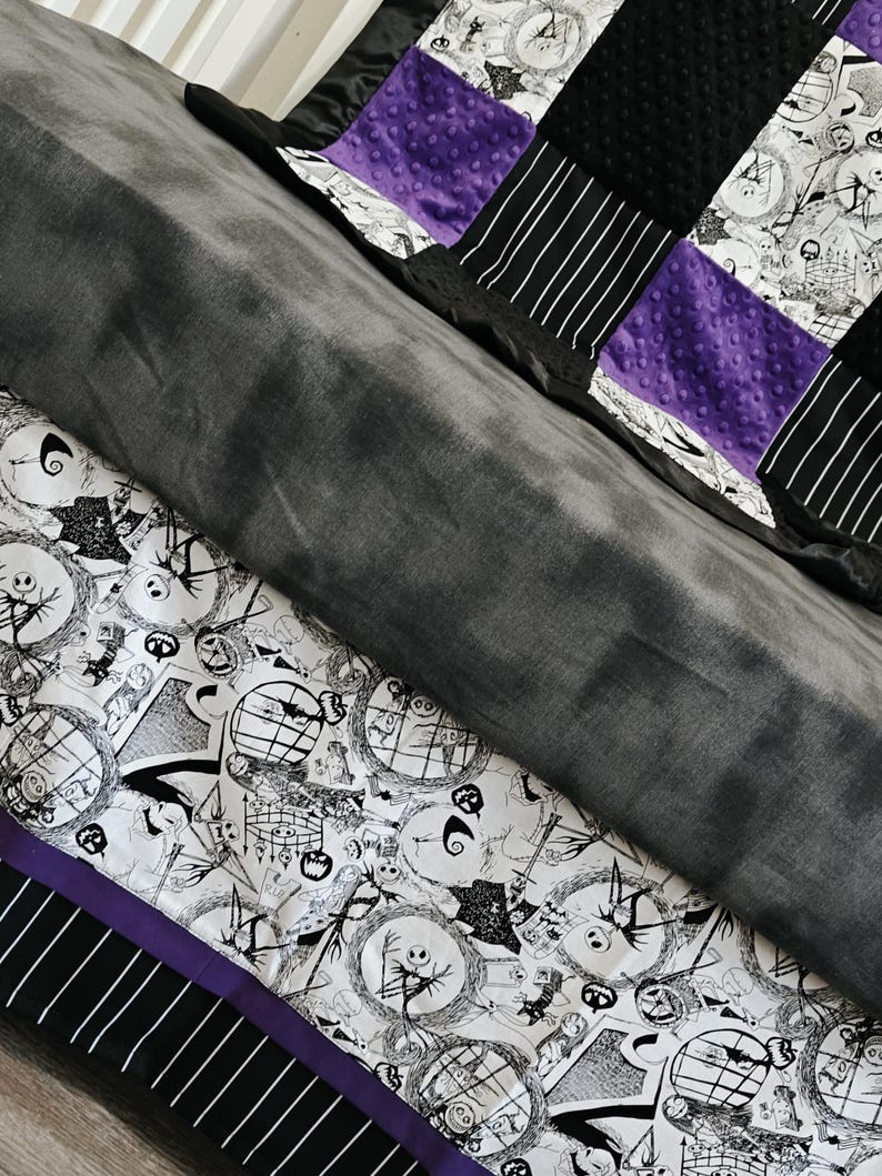 May include: A patchwork quilt featuring a black and white print with Jack Skellington characters, black and white striped panels, and purple accents. The quilt has a soft, gray minky fabric on the back.