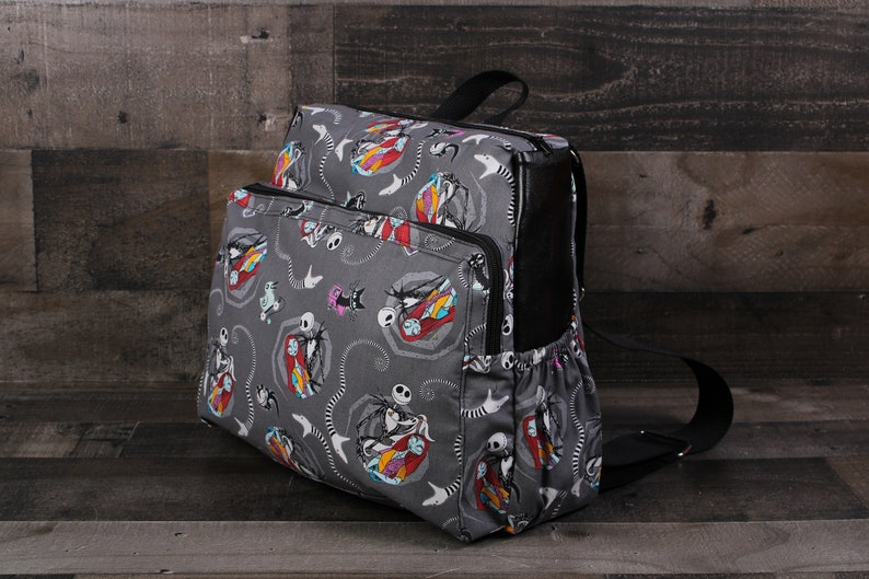 May include: A gray backpack with a black zipper and straps. The backpack has a repeating pattern of Jack Skellington and Sally from the Nightmare Before Christmas.
