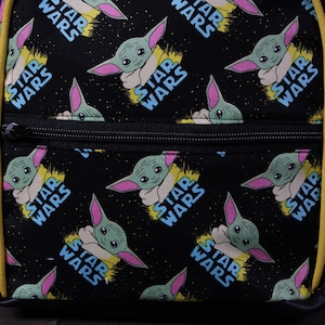 Puede incluir: Una mochila negra con ribete amarillo y un patr&oacute;n repetido de un personaje alien&iacute;gena verde con las palabras "Star Wars" sobre un fondo negro.