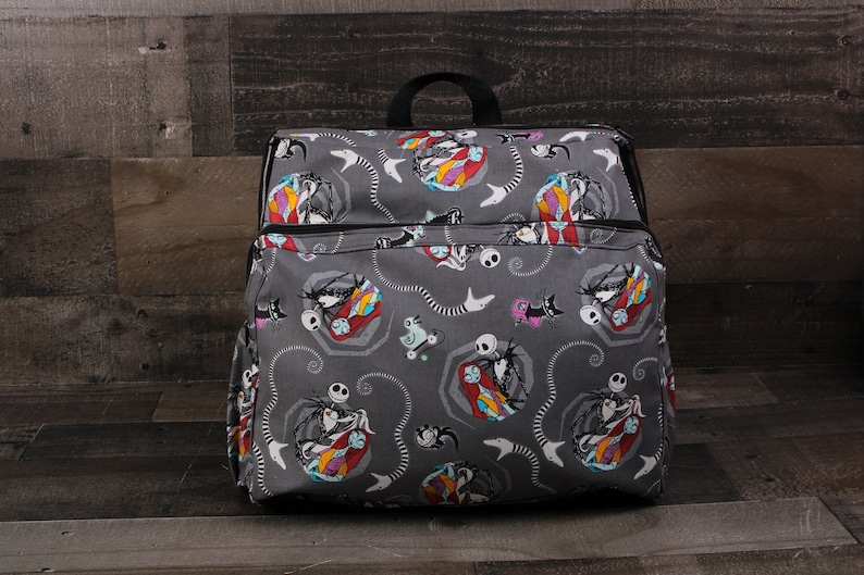 May include: A gray canvas backpack with a repeating pattern of Jack Skellington and Sally from The Nightmare Before Christmas. The backpack has a black zipper and black straps.