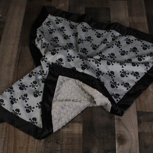 May include: A soft, gray and white baby blanket with a black skull and crossbones pattern. The blanket has a black satin border and a soft, white minky backing.
