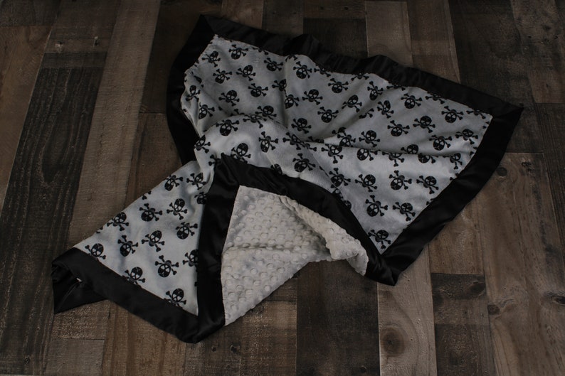 May include: A soft, gray and white baby blanket with a black skull and crossbones pattern. The blanket has a black satin border and a soft, white minky backing.