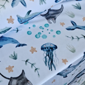 May include: White fabric with a watercolor pattern of marine life. The design includes blue dolphins, gray manta rays, blue jellyfish, and tan starfish. Green seaweed and teal bubbles complete the ocean-themed print.