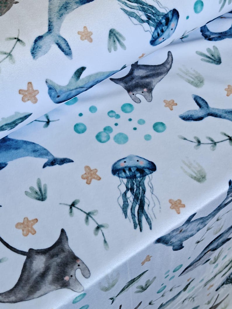 May include: White fabric with a watercolor pattern of marine life. The design includes blue dolphins, gray manta rays, blue jellyfish, and tan starfish. Green seaweed and teal bubbles complete the ocean-themed print.