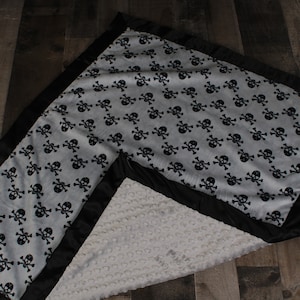 May include: A soft, gray and black baby blanket with a skull and crossbones pattern. The blanket has a black satin border and a soft, white, textured back.