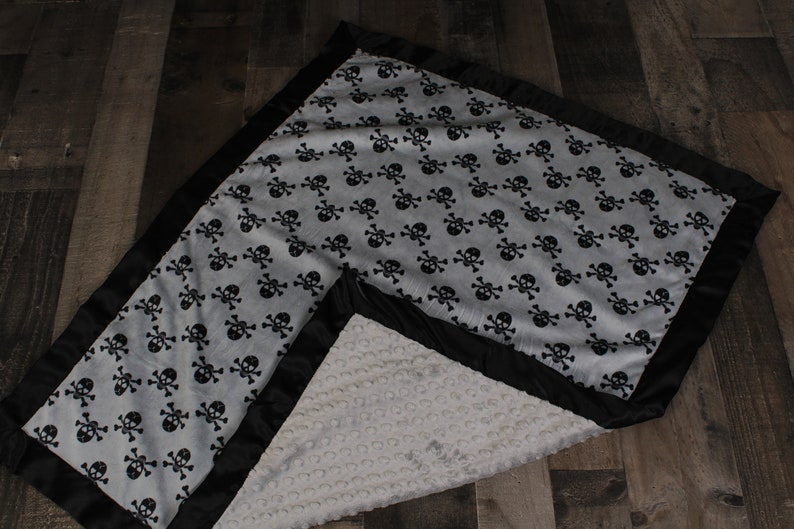 May include: A soft, gray and black baby blanket with a skull and crossbones pattern. The blanket has a black satin border and a soft, white, textured back.