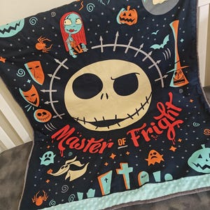 Puede incluir: Una manta azul oscuro con una gran cara de Jack Skellington y las palabras "Master of Fright". La manta tiene varias ilustraciones tem&aacute;ticas de Halloween, incluyendo a Sally, calabazas y fantasmas. El borde es azul claro.