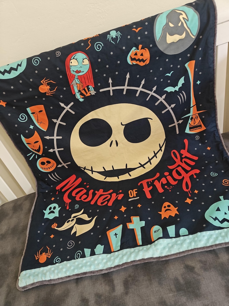 Puede incluir: Una manta azul oscuro con una gran cara de Jack Skellington y las palabras "Master of Fright". La manta tiene varias ilustraciones tem&aacute;ticas de Halloween, incluyendo a Sally, calabazas y fantasmas. El borde es azul claro.