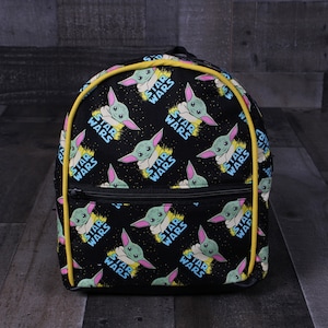 Puede incluir: Mochila negra con ribete amarillo y un patr&oacute;n repetido del personaje "Baby Yoda" de Star Wars. El patr&oacute;n tambi&eacute;n incluye las palabras "Star Wars" repetidas a lo largo.