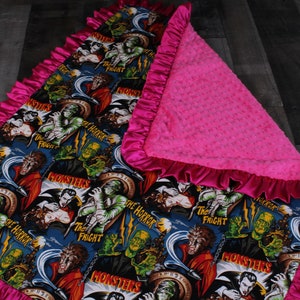 May include: A pink and blue baby blanket with a monster theme. The front of the blanket features a repeating pattern of classic monster movie characters, including Frankenstein, Dracula, and the Wolfman. The back of the blanket is a soft, pink minky fabric.