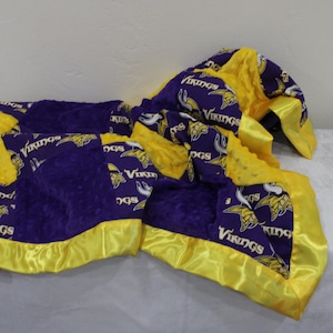 May include: A purple and yellow fleece blanket with a satin trim. The blanket features a repeated pattern of the Minnesota Vikings logo.