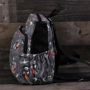 May include: A gray backpack with a black strap and a colorful print of characters from the Nightmare Before Christmas. The backpack has a zippered pocket on the front and a small pocket on the side.
