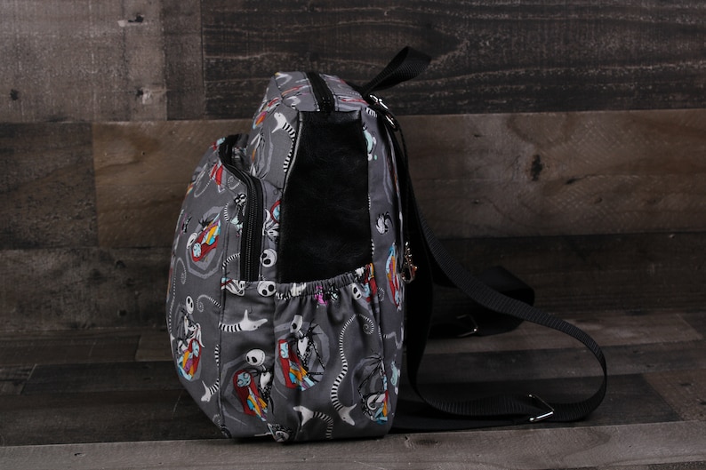 May include: A gray backpack with a black strap and a colorful print of characters from the Nightmare Before Christmas. The backpack has a zippered pocket on the front and a small pocket on the side.