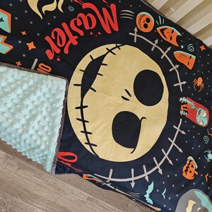 Puede incluir: Una manta para beb&eacute; negra y verde azulado con una gran cara de Jack Skellington y la palabra "Master" en rojo. La manta tiene un reverso texturizado verde azulado y un ribete gris. Tem&aacute;tica de Halloween.