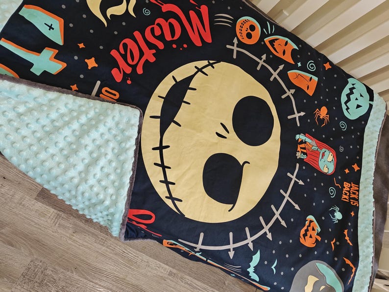 Puede incluir: Una manta para beb&eacute; negra y verde azulado con una gran cara de Jack Skellington y la palabra "Master" en rojo. La manta tiene un reverso texturizado verde azulado y un ribete gris. Tem&aacute;tica de Halloween.