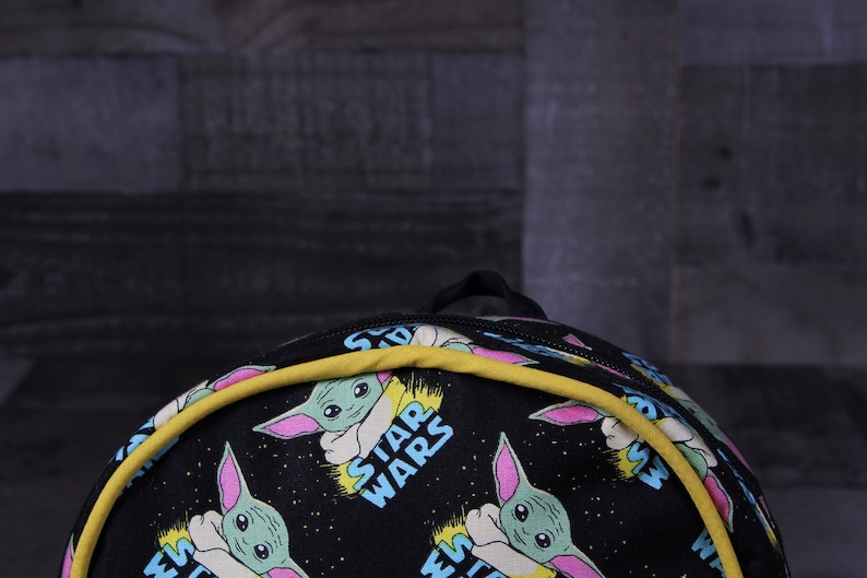 Puede incluir: Mochila negra con ribete amarillo y un patr&oacute;n repetido de un personaje verde, rosa y azul con el texto "Star Wars" sobre un fondo negro.