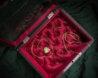 May include: A red velvet-lined jewelry box with a gold heart-shaped locket and several gold rings. The box is open and the jewelry is visible.