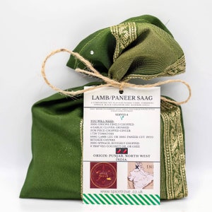 May include: A green fabric bag with a gold trim tied with twine. A white tag is attached with the text "LAMB/PANEER SAAG" and a list of ingredients needed to make the dish. The tag also includes the text "ORIGIN: PUNJAB, NORTH WEST INDIA."