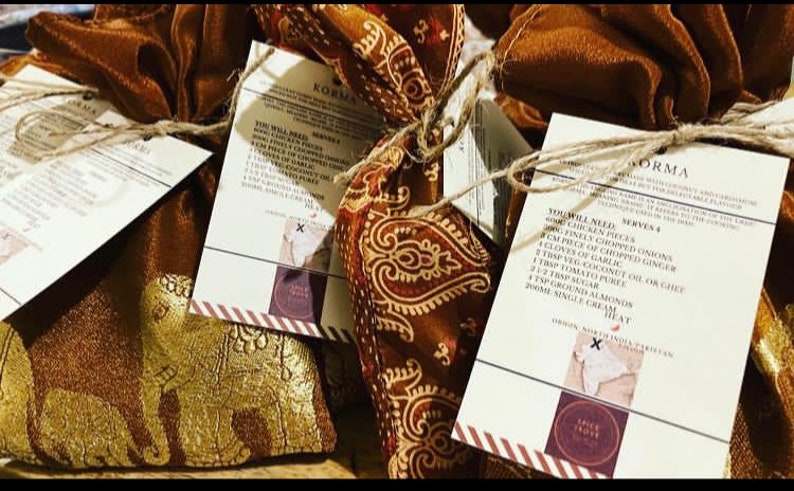 Peut inclure: Quatre petites pochettes en tissu brun avec des broderies dor&eacute;es, attach&eacute;es avec de la ficelle et &eacute;tiquet&eacute;es avec des cartes de recettes pour un plat Korma. Les cartes de recettes &eacute;num&egrave;rent les ingr&eacute;dients et les instructions pour pr&eacute;parer le plat. Les cartes ont une bordure ray&eacute;e rouge et blanche et un logo avec les mots "Spice Store" dans un cercle.