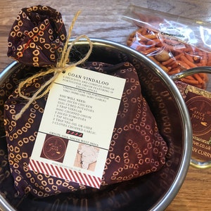 May include: A brown fabric pouch tied with twine and labelled "Goan Vindaloo" with a list of ingredients. The pouch is in a silver bowl with a bag of orange and red crunchy snacks. The label also includes the text "Spice Trove" and a map of India.