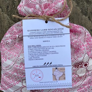 May include: A pink and white patterned fabric bag tied with twine, containing a recipe card for Kashmiri Lamb Rogan Josh. The card includes a list of ingredients and instructions for making the dish, along with a map of India highlighting the origin of the dish.