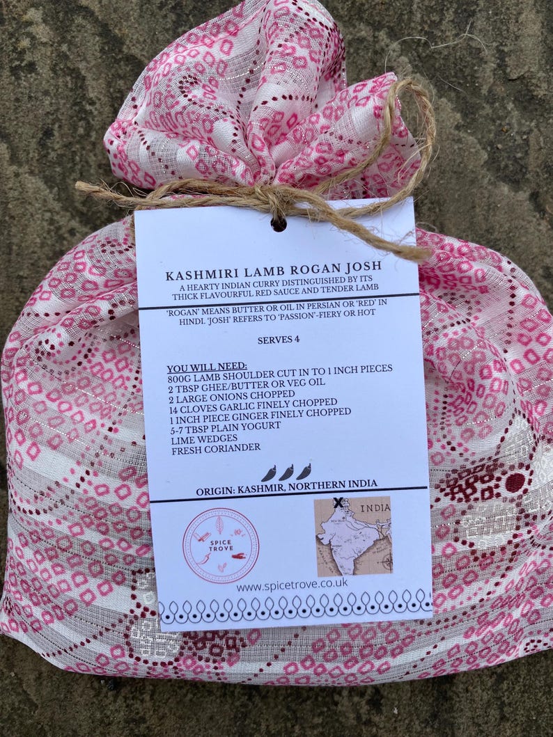 May include: A pink and white patterned fabric bag tied with twine, containing a recipe card for Kashmiri Lamb Rogan Josh. The card includes a list of ingredients and instructions for making the dish, along with a map of India highlighting the origin of the dish.