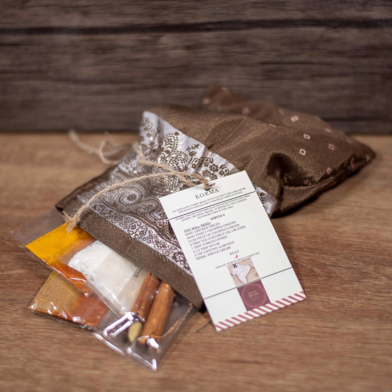 Peut inclure: Une pochette en tissu marron &agrave; motif cachemire nou&eacute;e avec de la ficelle. La pochette contient plusieurs petits sachets d'&eacute;pices et une cannelle. Une carte blanche avec le texte "KORMA" et une recette de Korma est attach&eacute;e &agrave; la pochette.