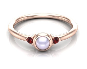 May include: A rose gold ring featuring a central round white pearl in a bezel setting, flanked by two small round red garnet gemstones on either side. The band is smooth and polished, creating a simple and elegant design with a warm metallic finish.