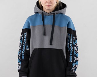 Puede incluir: Una sudadera con capucha de bloques de color que presenta capucha y pecho gris, sección media azul, y cuerpo inferior y mangas negras. Las mangas están decoradas con un patrón geométrico en azul, blanco y negro. Incluye bolsillo frontal y cordones negros.