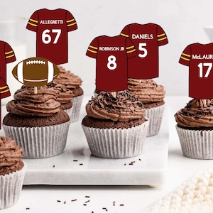 May include: Six chocolate cupcakes decorated with brown sprinkles and topped with football jersey themed cupcake toppers. The jerseys are burgundy with gold trim and feature the names and numbers of different football players. The football players' names are Cosmi, Allegretti, Robinson Jr, Daniels, McLaurin, and Ekeler.