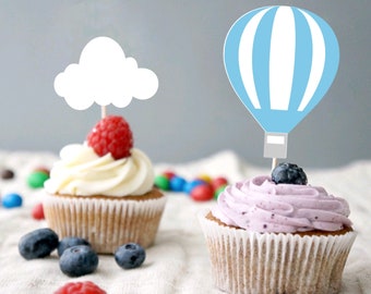 May include: Two cupcakes decorated with blue and white hot air balloon and white cloud toppers. One cupcake has white frosting and a red raspberry on top. The other cupcake has purple frosting and a blueberry on top. Both cupcakes are surrounded by blueberries and colorful candy.