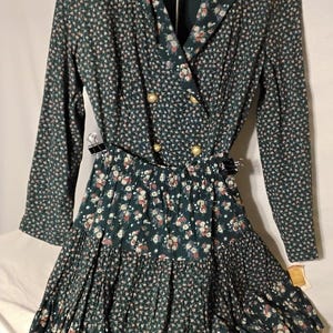 May include: A dark green long-sleeve dress featuring a small floral pattern in white, pink, and blue. The bodice has a notched lapel collar, four gold-toned buttons, and a drop-waist design. The skirt consists of two gathered, tiered ruffles.