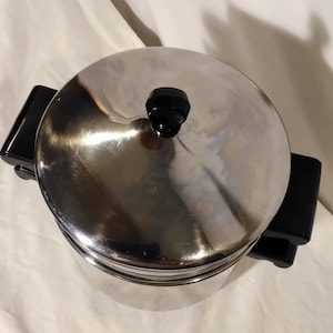 May include: A cylindrical stainless steel ice bucket with a polished silver finish. It features two black rectangular side handles and a matching black knob on the circular lid. The metal surface reflects light, showing a smooth, metallic texture.