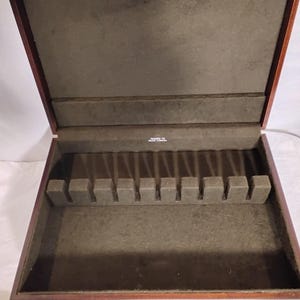 May include: Open wooden silverware storage box with a dark brown exterior and a green felt interior. The box has a rectangular shape with a hinged lid and a series of slots for holding utensils. The interior is lined with a soft, green felt material.