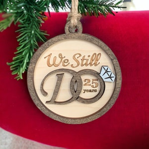 Peut inclure: Une décoration de Noël en bois avec le texte "We Still Do" et une bague avec un diamant. La bague porte le nombre 25 et le mot "years" inscrit dessus.