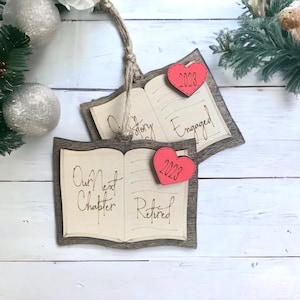 May include: Two wooden book-shaped ornaments with red hearts and the text "Our Next Chapter Retired 2023" and "Our Story Engaged 2023".