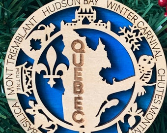 May include: A wooden Christmas ornament with a blue background, featuring a map of Quebec and surrounding landmarks. The ornament includes the text "QUEBEC" and other place names. It is designed with a circular shape and a hole for hanging.