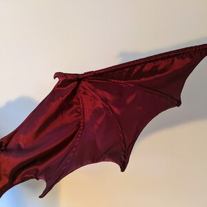 May include: A pair of red fabric wings with a textured surface. The wings are designed to look like a dragon's wings.
