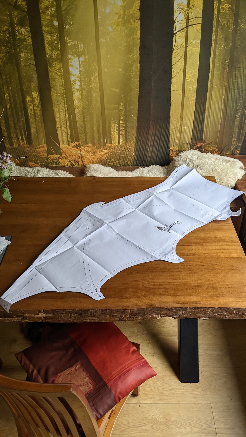 May include: A white paper pattern for a pair of wings, folded on a wooden table. The pattern is labeled "Camera Wings".