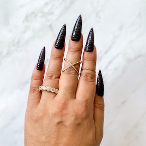 May include: A hand with long, black, pointed nails with a snakeskin pattern. The hand is wearing two gold rings, one with a cross design and one with a band of small diamonds.
