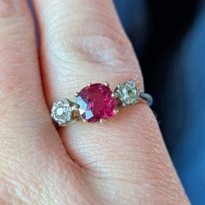 Peut inclure: Une bague en or ornée d'une pierre rouge vif de forme ovale au centre, flanquée de deux petites pierres rondes et transparentes. La bague a un anneau fin et les pierres sont serties dans un style classique.