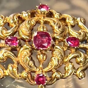 May include: Ornate gold-toned brooch featuring a central, large, ruby-red gemstone, surrounded by smaller ruby-red stones. The brooch has a detailed, swirling, and floral design. The overall aesthetic is vintage and luxurious.