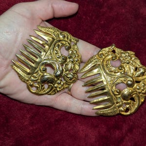 May include: Two ornate, gold-colored hair combs with intricate floral and scroll designs. The combs are held in a hand, showcasing their detailed craftsmanship and vintage aesthetic. The background is a deep red fabric.