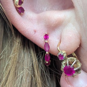 May include: Close-up of an ear adorned with multiple pieces of jewelry. A gold stud earring with a heart-shaped design and a ruby stone is visible. A small gold hoop earring with three ruby stones is also present, along with a gold earring with a ruby stone.