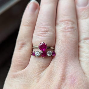 May include: Two silver rings with gemstones. The top ring features a large, oval-cut ruby. The bottom ring has three rubies and two smaller, clear stones. The rings are worn on a hand.