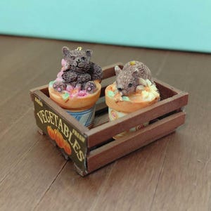 May include: Miniature figurines of a bear and a mouse atop decorative ice cream cones, nestled in a small wooden crate. The crate has the words "Fresh Vegetables" printed on the side. The figurines are detailed and colorful, with a whimsical aesthetic.
