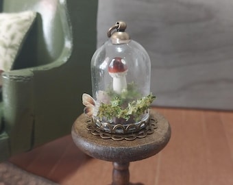 May include: A miniature glass cloche with a mushroom inside, set on a small wooden table. The mushroom has a red and white cap and sits among green moss. A decorative butterfly is attached to the base of the cloche. The table has a dark brown finish.